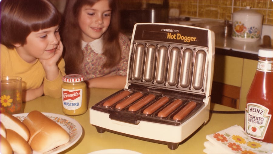Two children look at a Presto Hot Dogger cooking six hot dogs on a kitchen table with mustard, ketchup, and hot dog buns nearby.