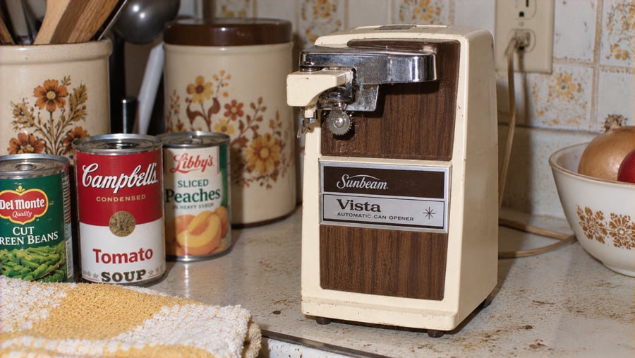 A vintage Sunbeam Vista electric can opener sits on a kitchen counter next to cans of green beans, tomato soup, and sliced peaches.