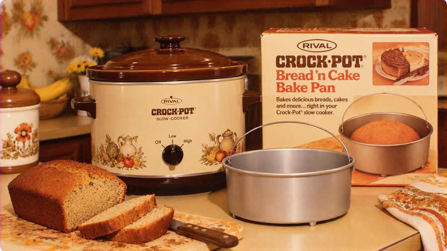 A Rival Crock-Pot slow cooker next to its original bread and cake bake pan in a box, with a sliced loaf of bread on a cutting board.