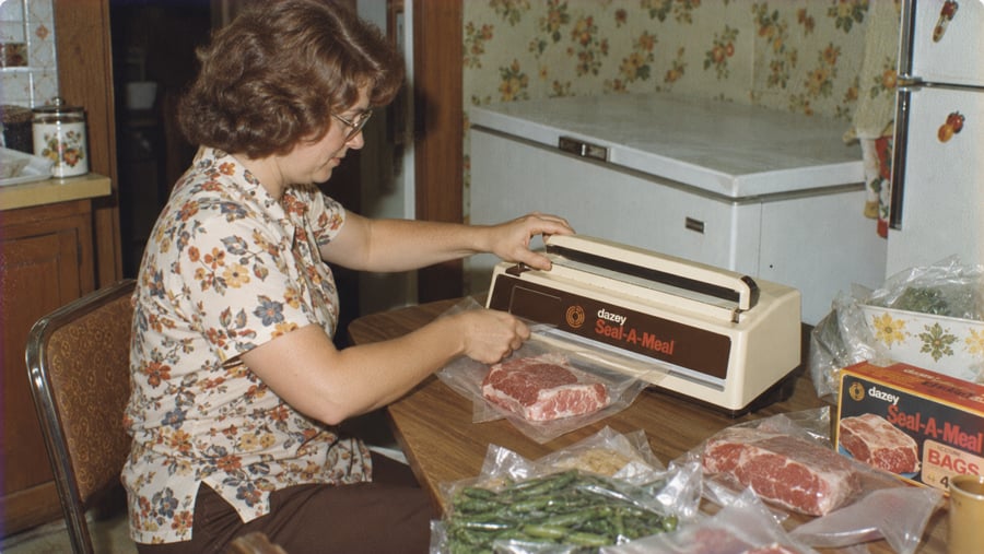 A woman uses a Dazey Seal-A-Meal machine to vacuum-seal pieces of raw meat on a kitchen table.