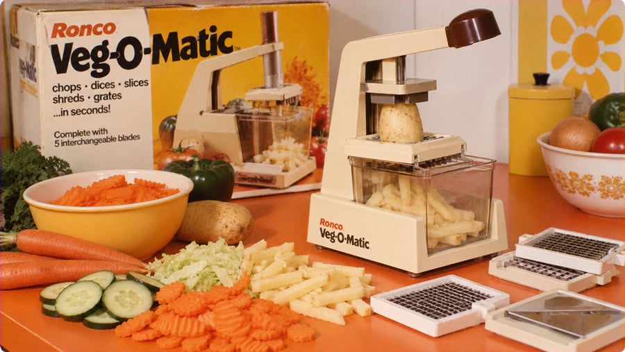 A Ronco Veg-O-Matic food slicer on a kitchen counter with interchangeable blades, surrounded by sliced carrots, cucumbers, and potatoes.