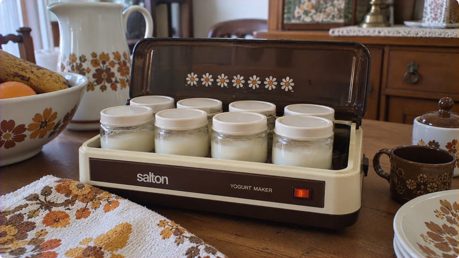 A vintage Salton yogurt maker with eight glass jars inside sits on a wooden table surrounded by matching floral-patterned kitchenware.