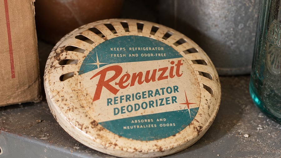 A vintage Renuzit refrigerator deodorizer with a round, vented lid sits on a shelf. The label is teal, red, and cream, and reads “Keeps refrigerator fresh and odor-free. Absorbs and neutralizes odors.”