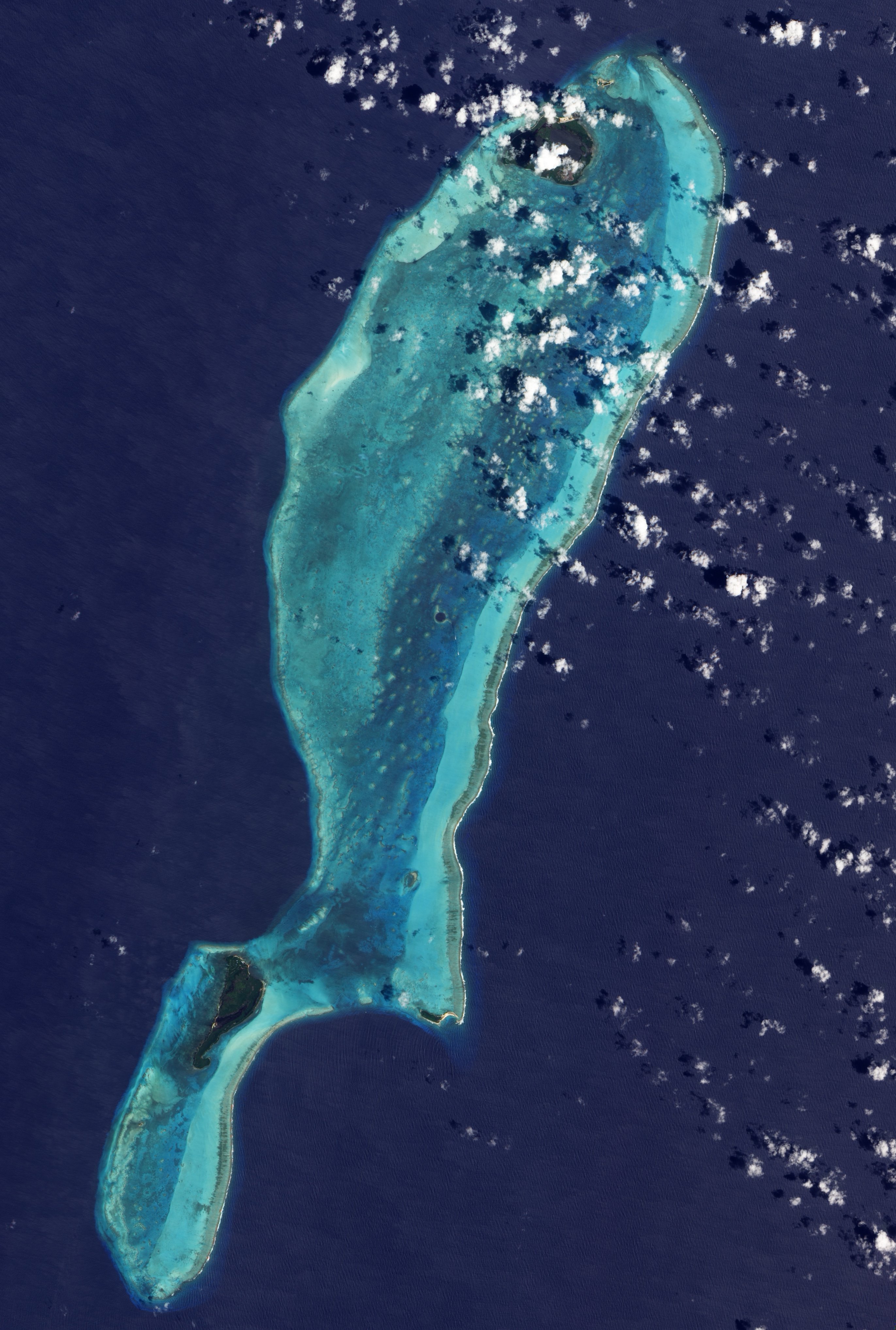 Satellite view of a long, narrow coral reef surrounded by deep blue ocean, with small islands at the north and south ends and scattered clouds overhead.