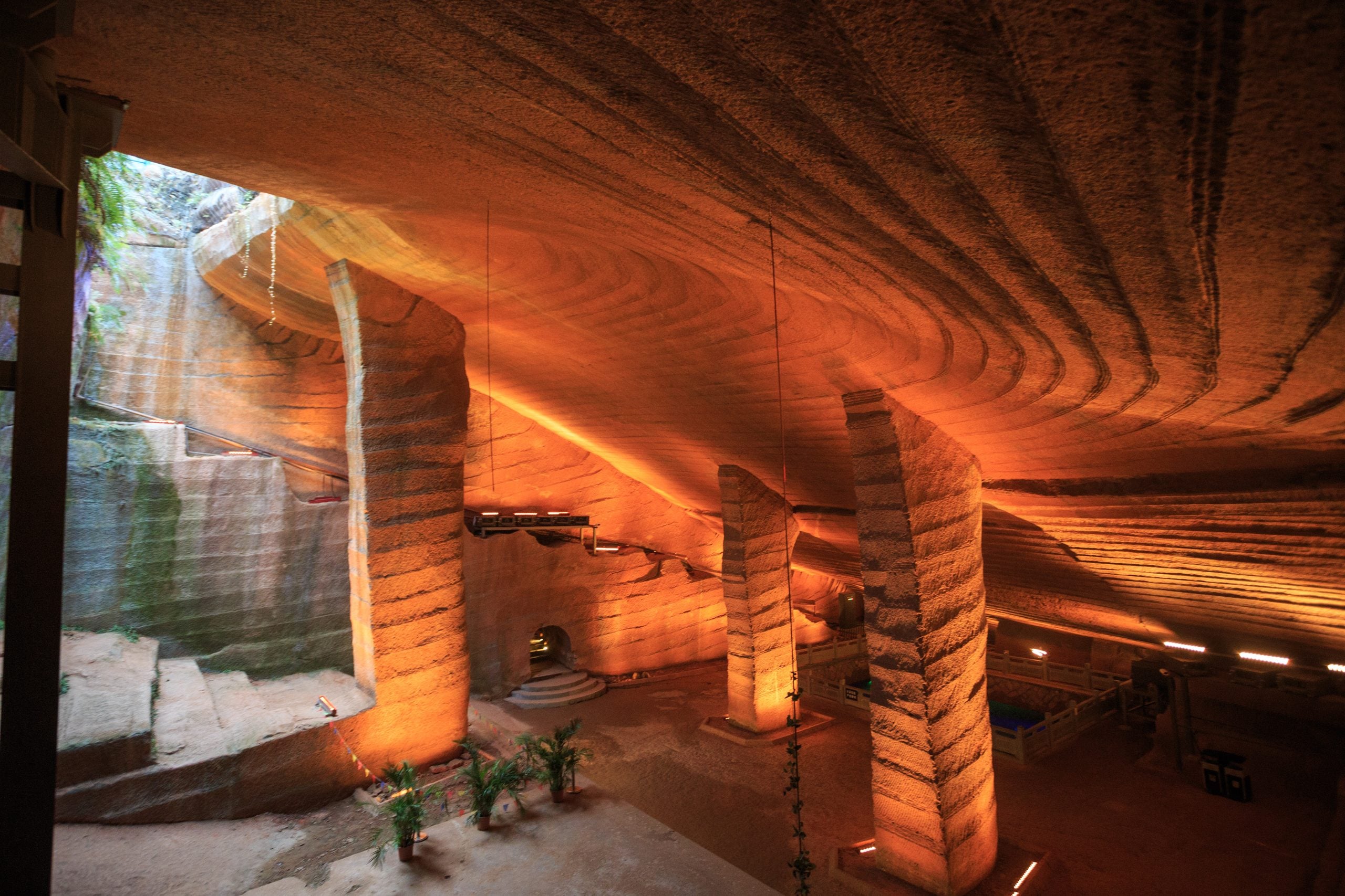 A massive man-made stone cavern with high, tiered ceilings and large carved pillars, illuminated by warm orange lighting.