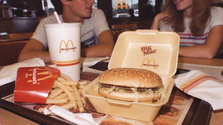 A tray with a McDonald’s Big Mac in a foam container, a carton of french fries, and a large drink cup, with two people sitting at a table in the background.