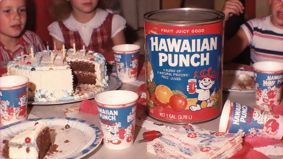 A birthday party table featuring a large gallon can of Hawaiian Punch, a chocolate cake with candles, and matching Hawaiian Punch paper cups.