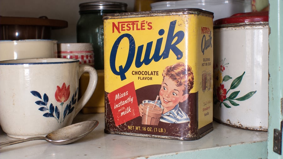 Vintage yellow Nestle’s Quik chocolate drink mix tin sitting on a shelf among assorted kitchenware.