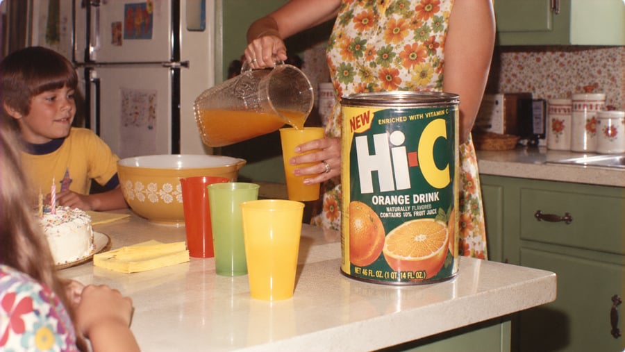 A person in a floral dress pours Hi-C orange drink from a pitcher into a plastic cup on a kitchen counter, where a young boy waits by a birthday cake.