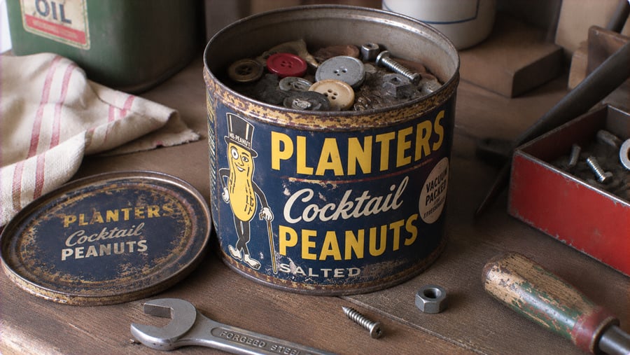 A vintage Planters Cocktail Peanuts tin filled with assorted buttons and hardware, sitting on a wooden workbench with a wrench and scattered screws.
