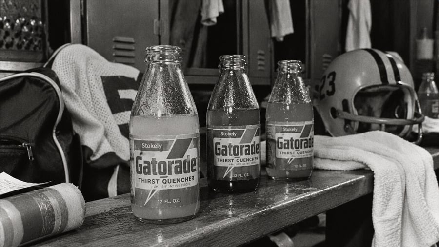 Three glass bottles of Gatorade on a wooden locker room bench near a football helmet and a sports bag.