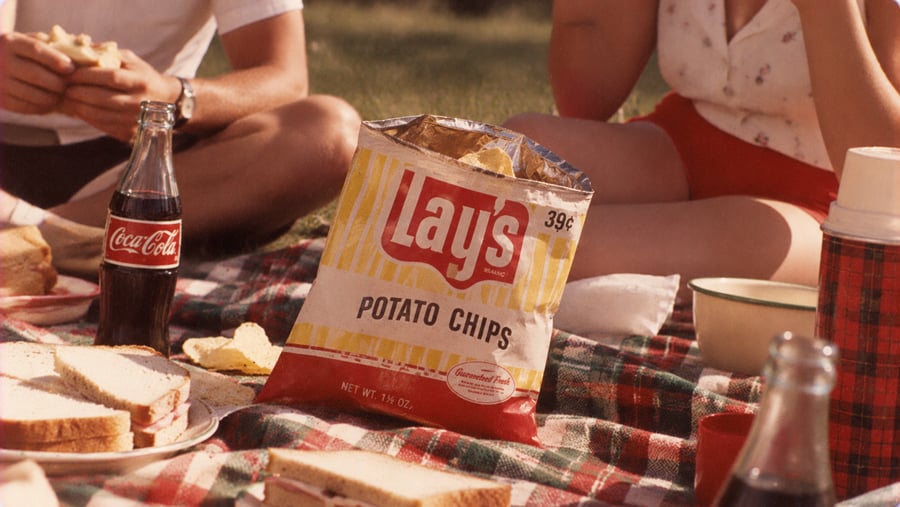 A picnic scene featuring a bag of Lay’s potato chips, a glass bottle of Coca-Cola, and plates of sandwiches on a plaid blanket.