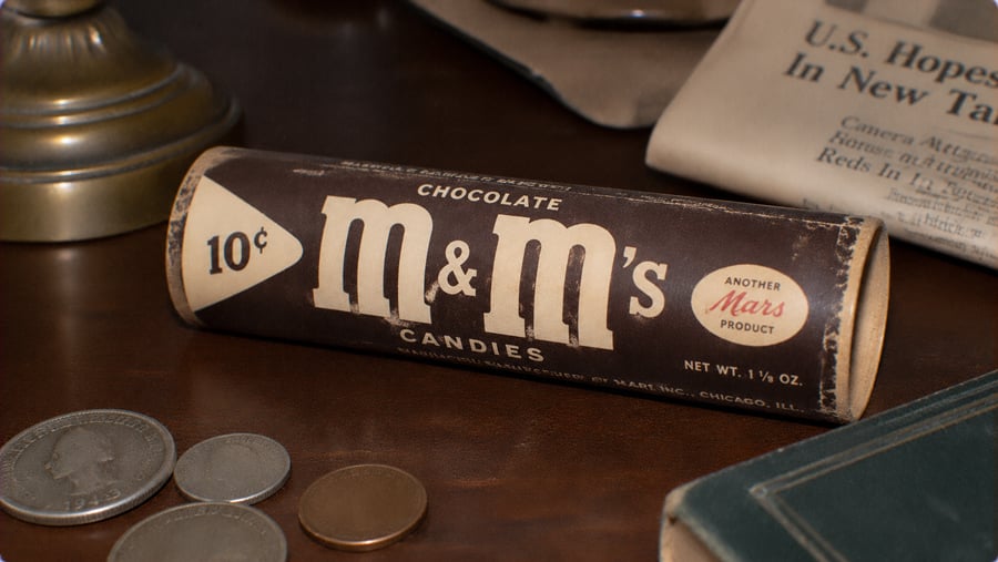 A vintage cylindrical cardboard package of M&M’s chocolate candies sitting on a wooden desk next to coins and a newspaper.