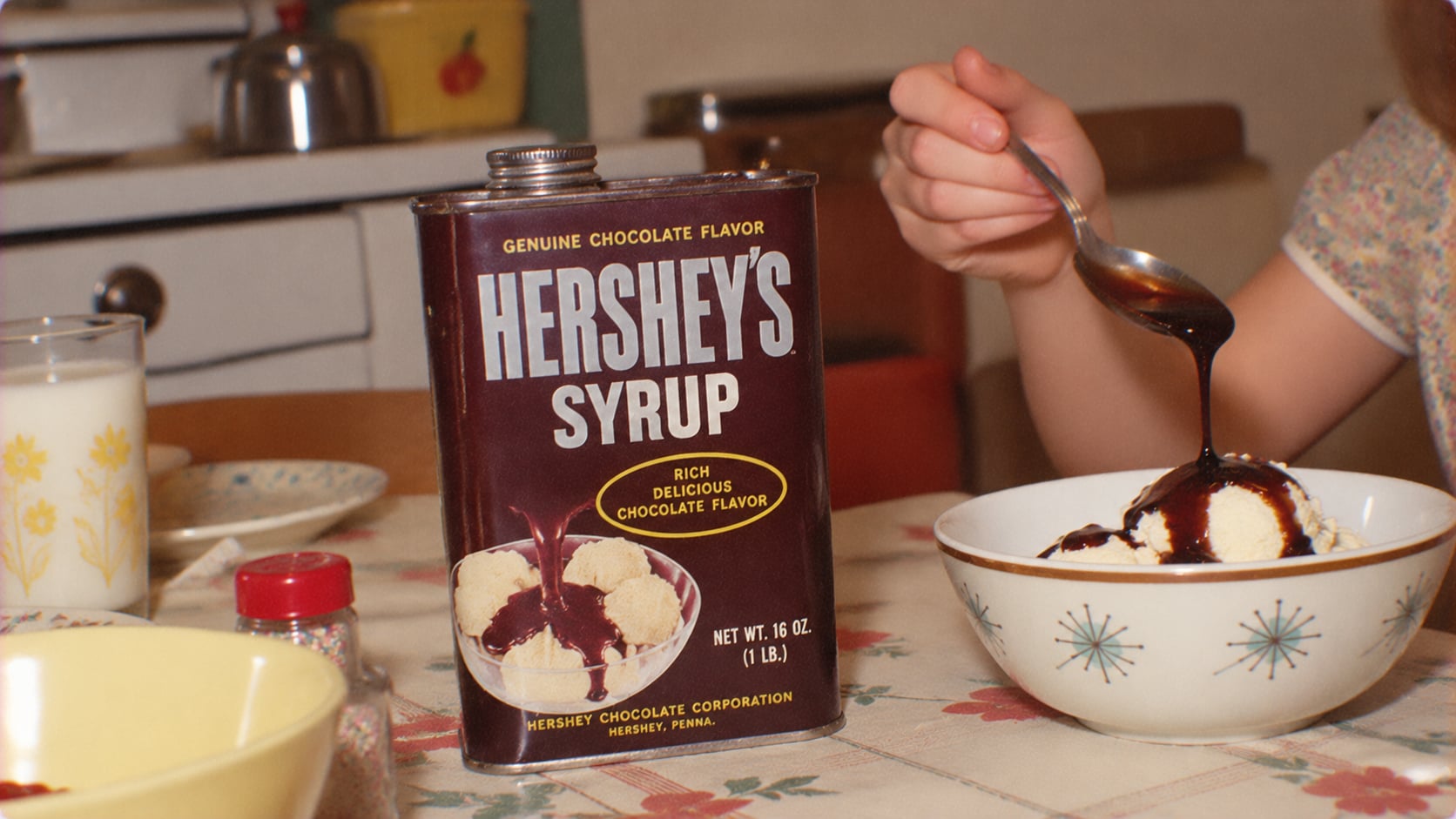 A vintage Hershey’s Syrup tin sits on a table next to a bowl of ice cream being drizzled with chocolate syrup by a hand holding a spoon.