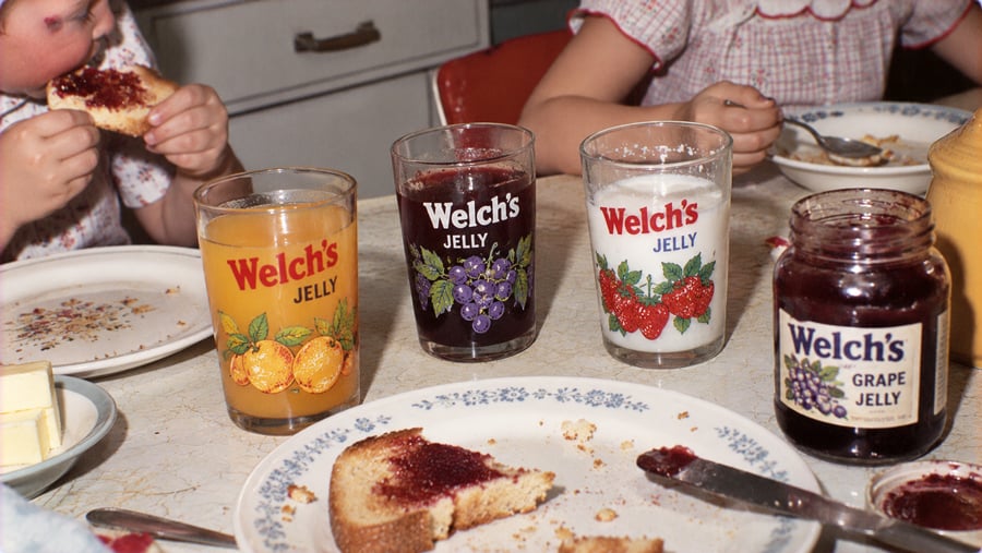 Children eat toast at a table set with three vintage Welch’s jelly glasses filled with juice, dark liquid, and milk, alongside an open jar of grape jelly.