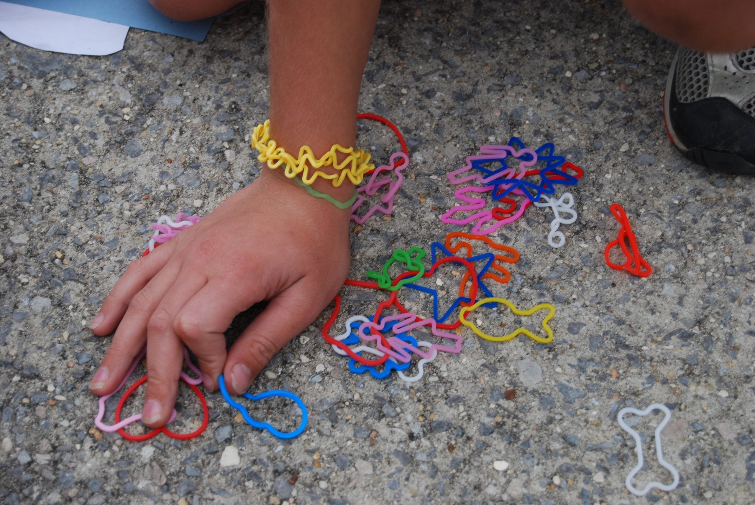 A hand resting on a gravel surface surrounded by various colorful, shaped rubber bands, with one yellow rubber band worn as a bracelet.