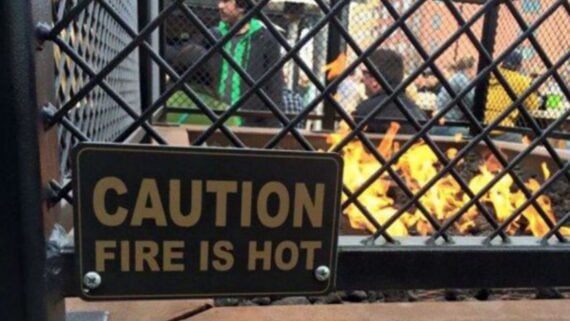 A “Caution: Fire is hot” sign attached to a metal fence in front of a fire pit.