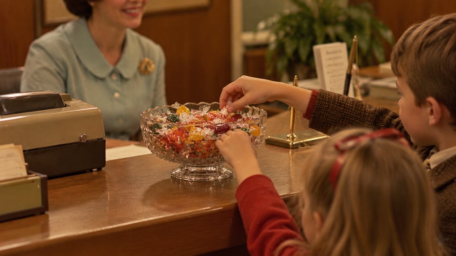 Two children reach into a glass bowl filled with assorted wrapped candies sitting on a wooden desk, while a woman in a blue dress sits behind them.