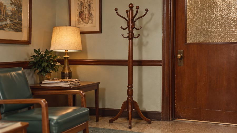 A wooden coat rack stands in the corner of an office next to a green armchair, a small side table with a lamp, and a wooden door.