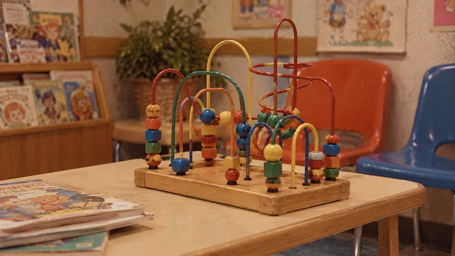 A wooden bead maze toy with colorful shapes sits on a table in a room with children’s books and plastic chairs.