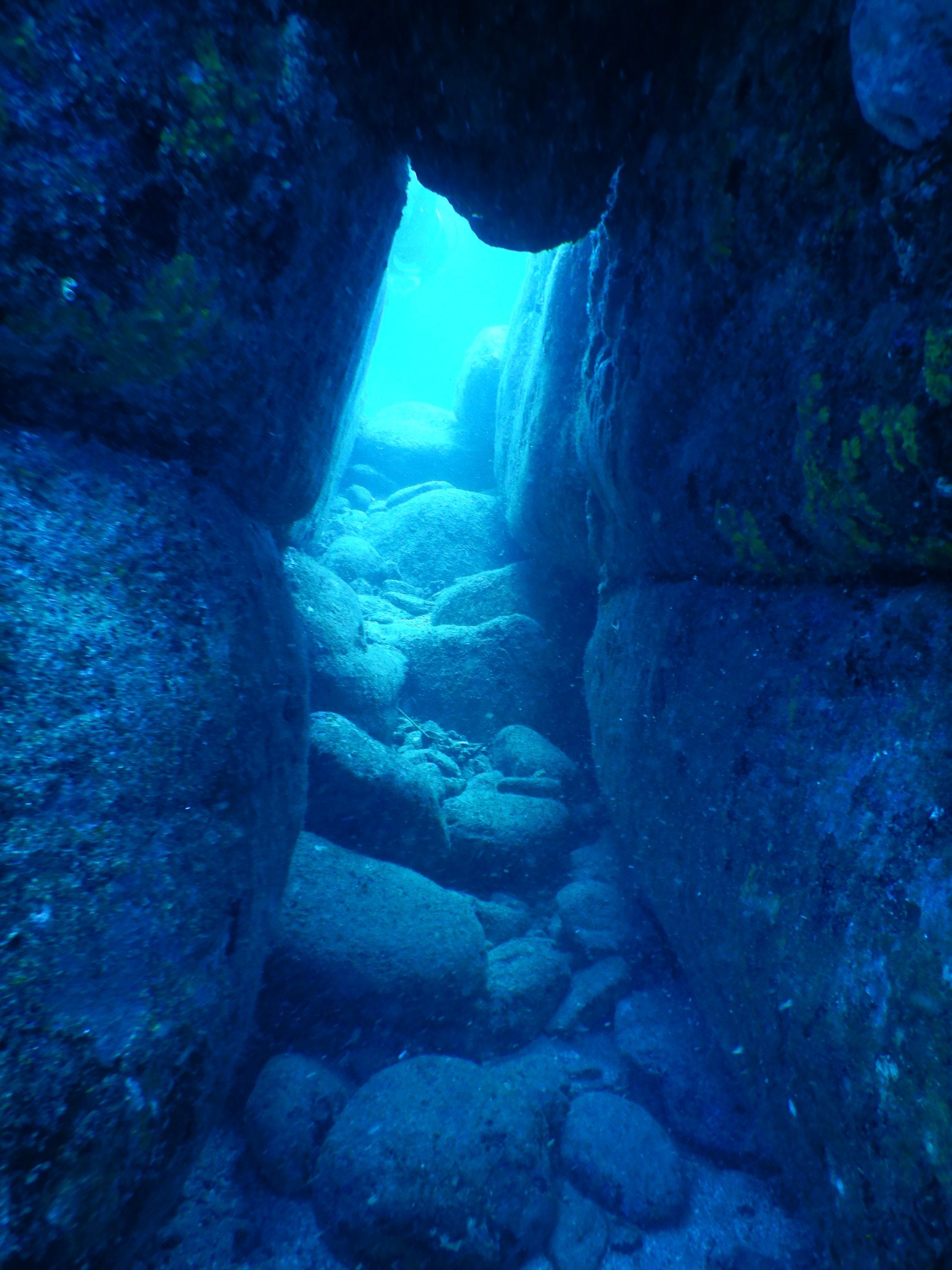 Underwater view of a narrow, light-filled rocky passage between large stone boulders.