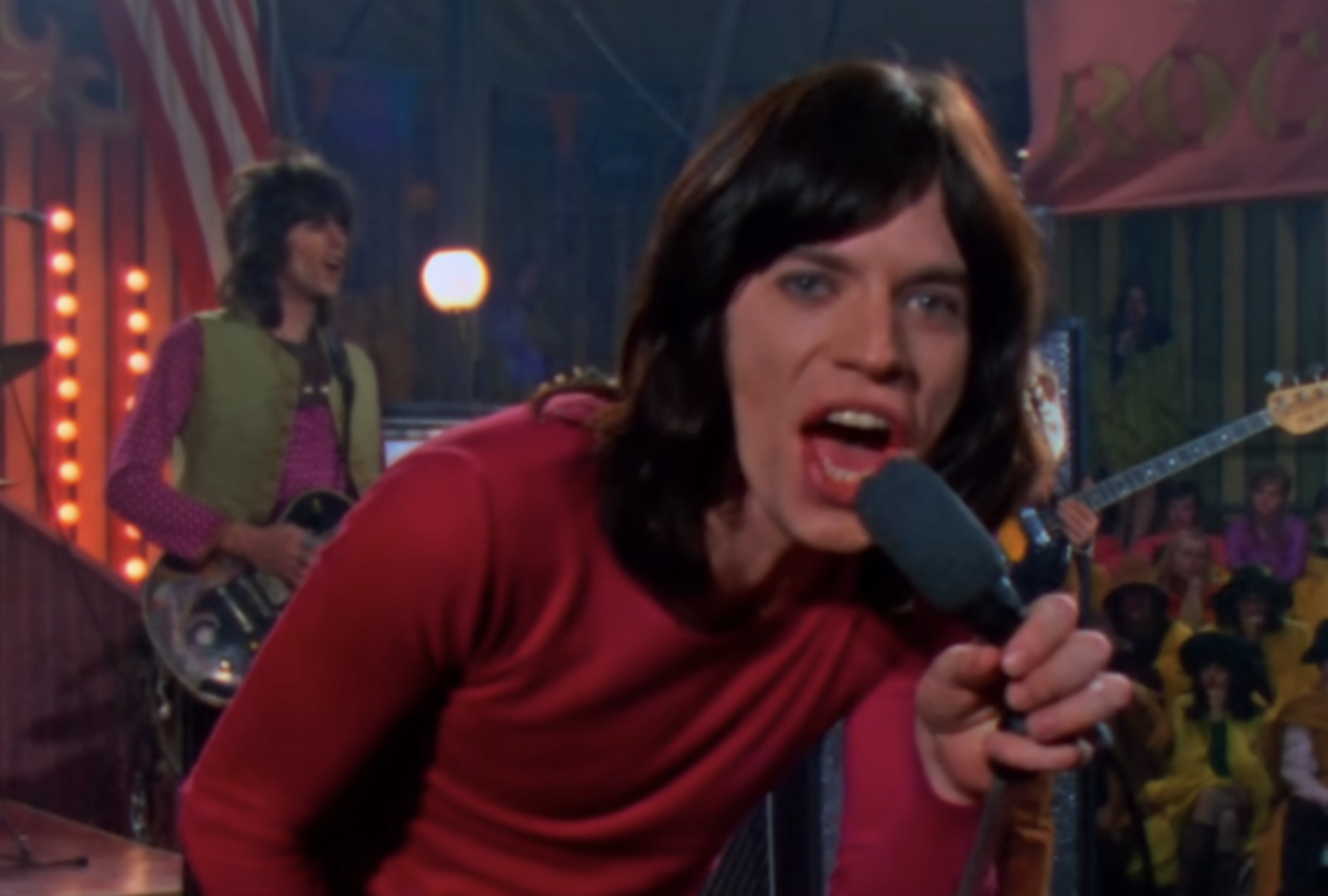 Mick Jagger singing into a microphone with Keith Richards playing guitar in the background.