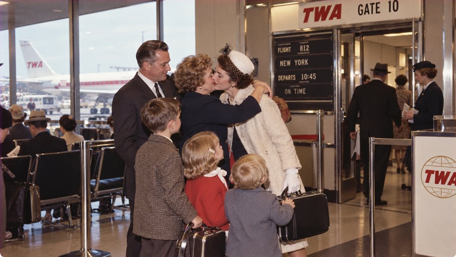 A woman in a pillbox hat kisses another woman goodbye at a TWA airport gate, observed by a man and two children holding suitcases.