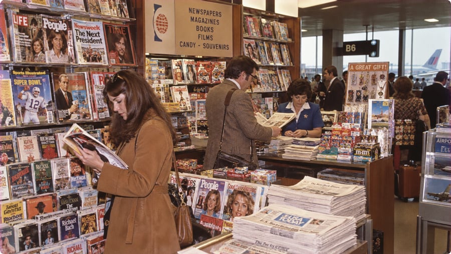 A woman browses magazines in an airport newsstand, with a cashier and other travelers in the background.