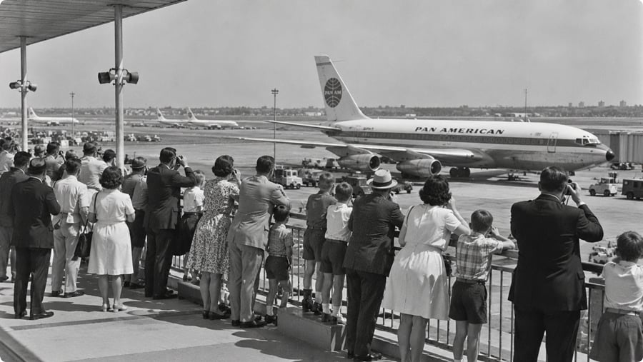 A crowd of adults and children stand on an observation deck watching a Pan American jet parked on the airport tarmac.