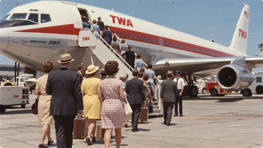 Passengers in mid-century attire board a TWA Boeing 707 airplane via an outdoor staircase on the tarmac.