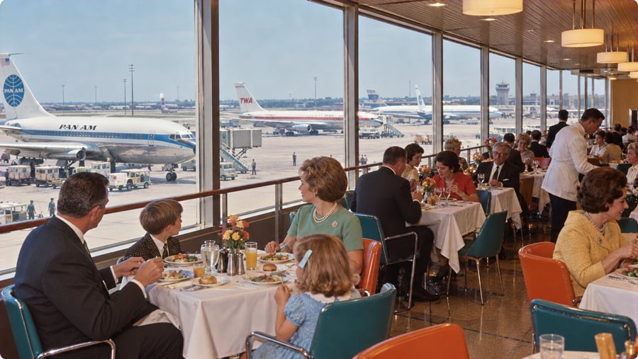 Diners eating at tables in an airport restaurant overlooking a tarmac with Pan Am and TWA airplanes.