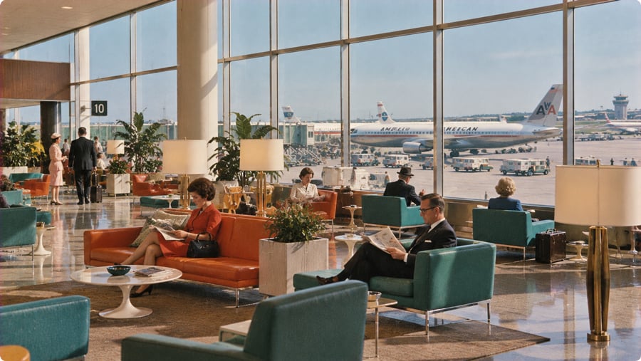 Travelers sit in a mid-century airport lounge with floor-to-ceiling windows overlooking American Airlines planes on the tarmac.