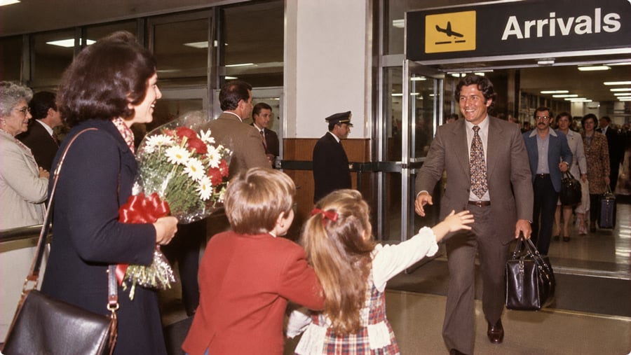 A man in a suit arriving at an airport terminal reaches out toward two children who are running to greet him, while a woman holding a bouquet of flowers stands nearby.