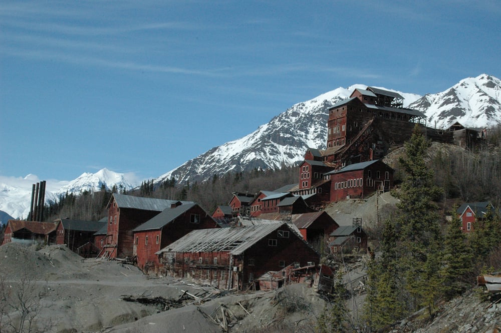 A collection of rustic red buildings from a historic mining site nestled at the base of snow-capped mountains.