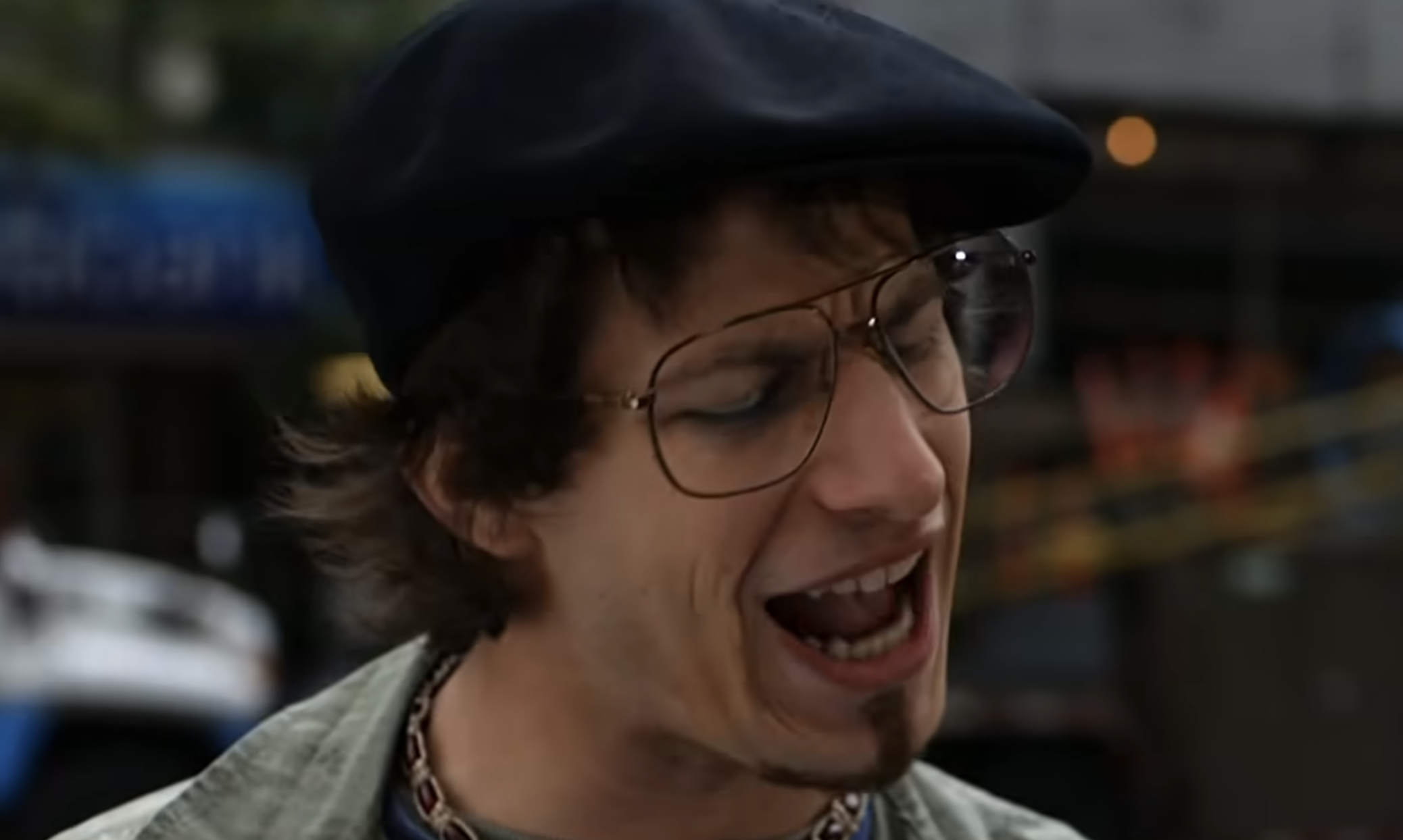 A man wearing a black flat cap and gold-rimmed glasses singing with his mouth open.