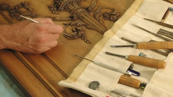 A woodworker uses a small metal tool to carve intricate detail into a wooden surface, with a rolled canvas kit of woodcarving tools laid out nearby.
