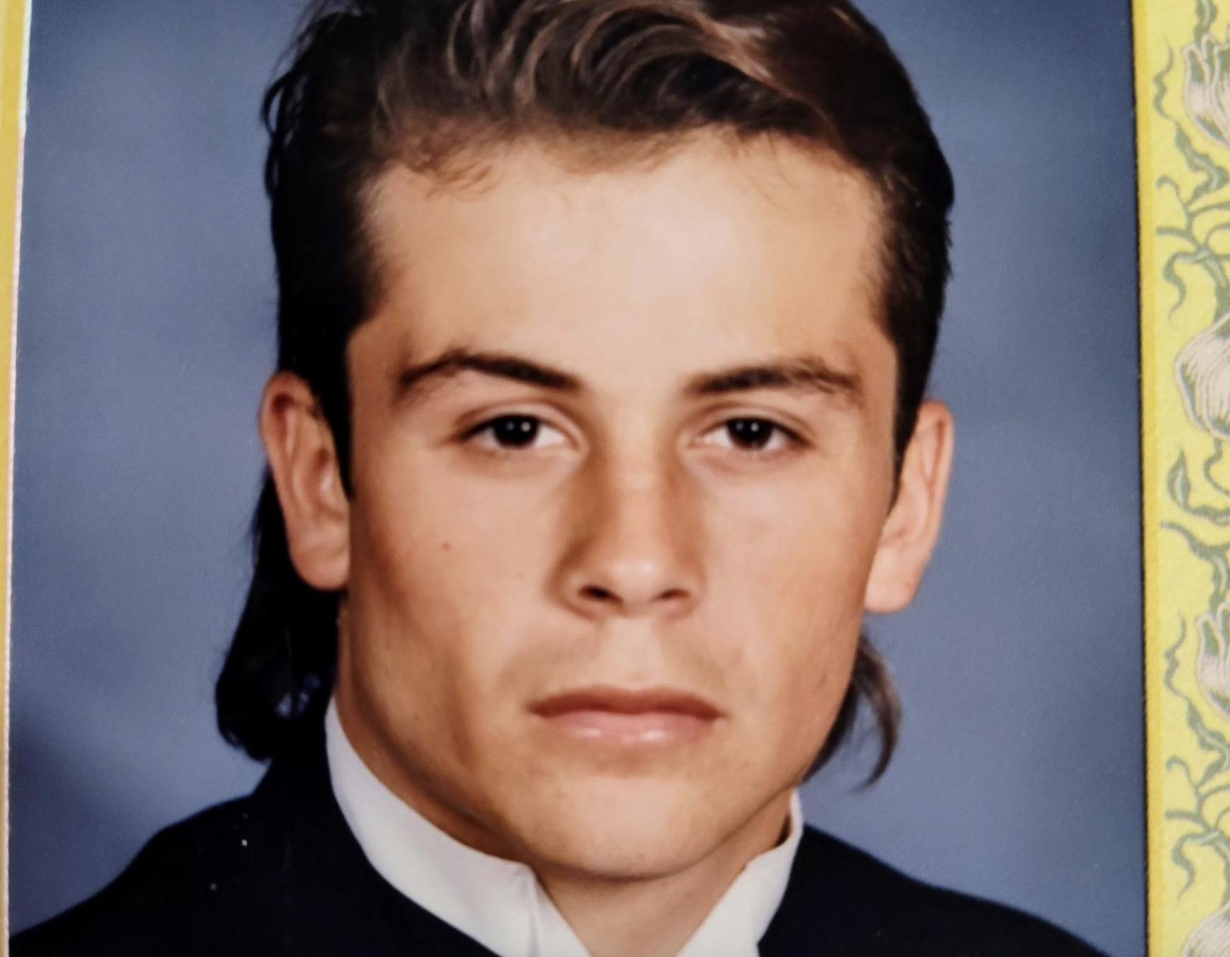 A headshot of a young man with a mullet hairstyle wearing a dark suit and white shirt against a blue background.