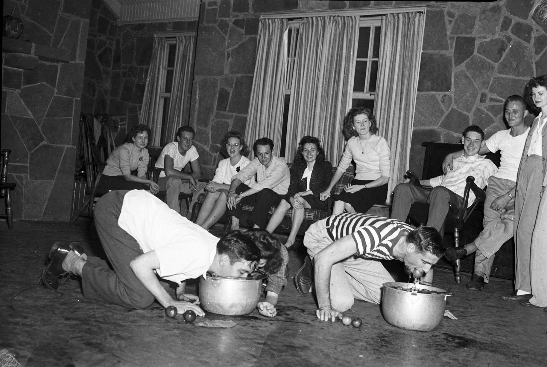 Two men on their knees on the floor bob for apples in metal basins while a group of people watches them.