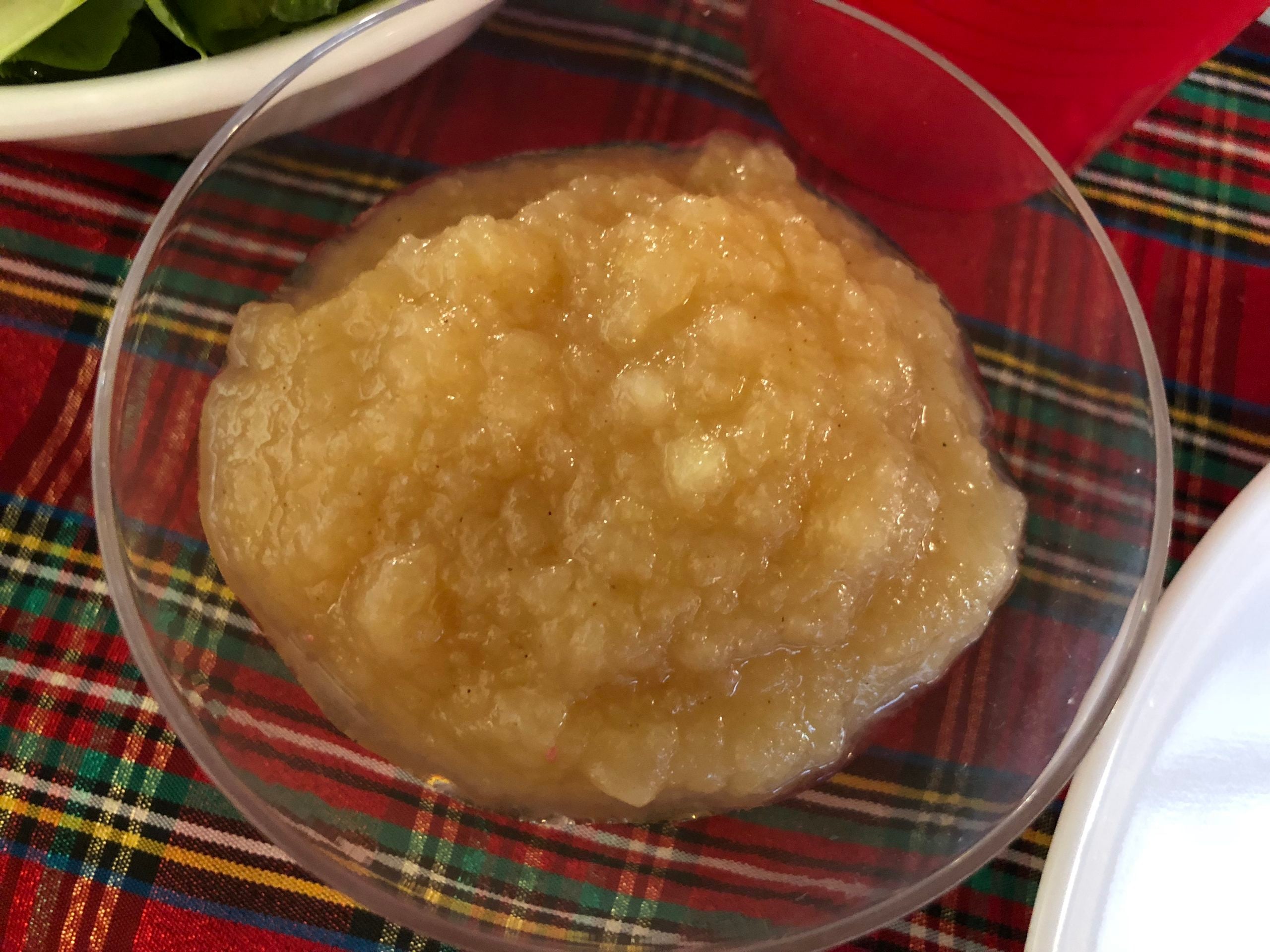 A clear bowl filled with applesauce sits on a red plaid tablecloth, next to a partially visible plate, cup, and greens.
