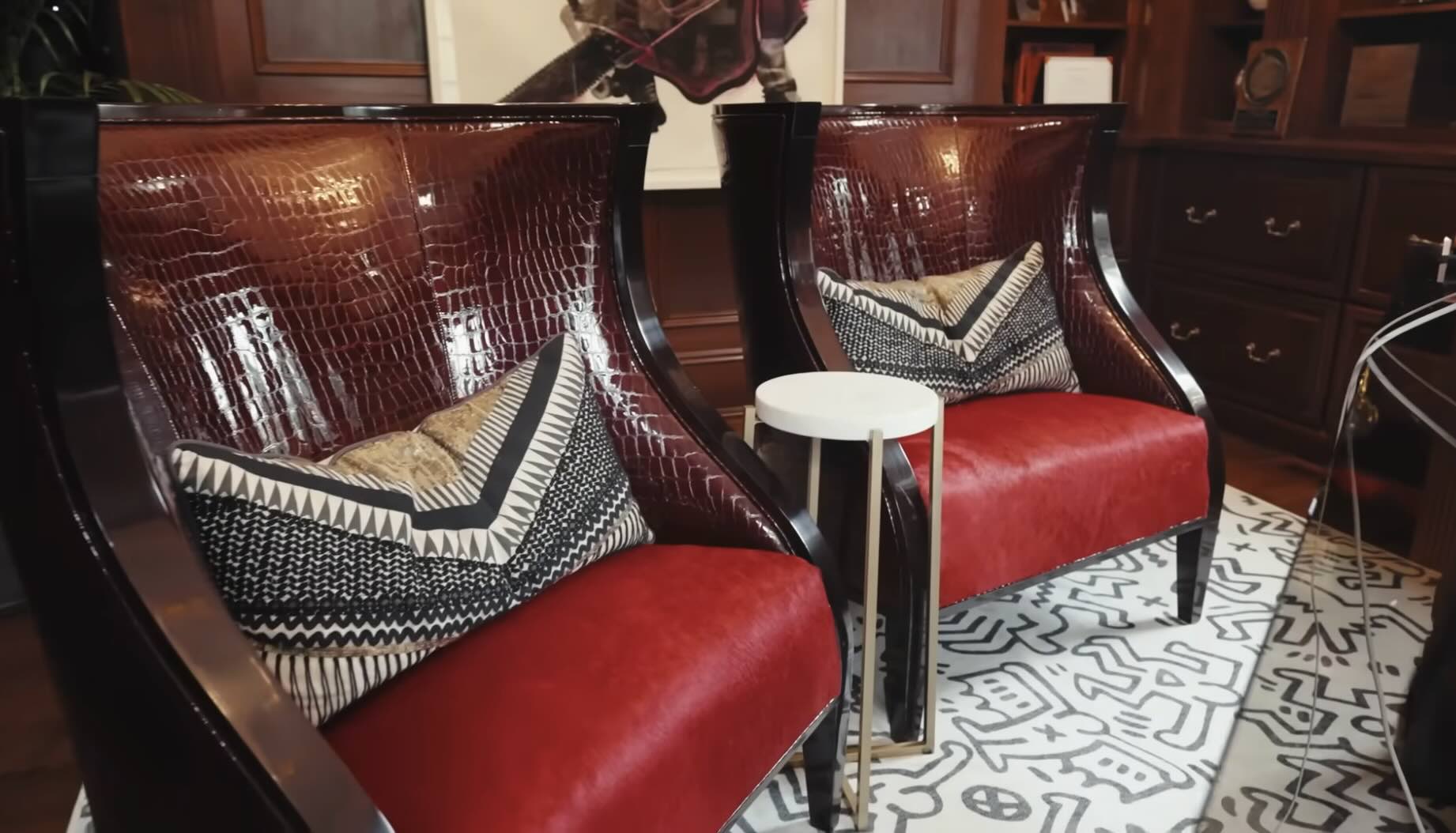 Two red armchairs with dark wood frames and patterned throw pillows, separated by a small white pedestal table on a geometric rug.
