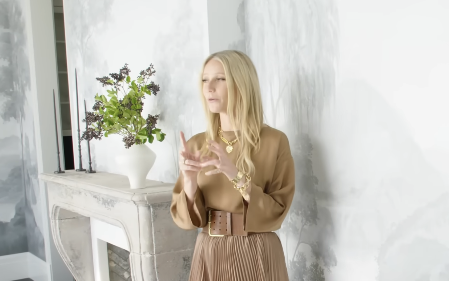 Gwyneth Paltrow wearing a tan blouse and pleated skirt, gesturing while speaking in front of a fireplace.