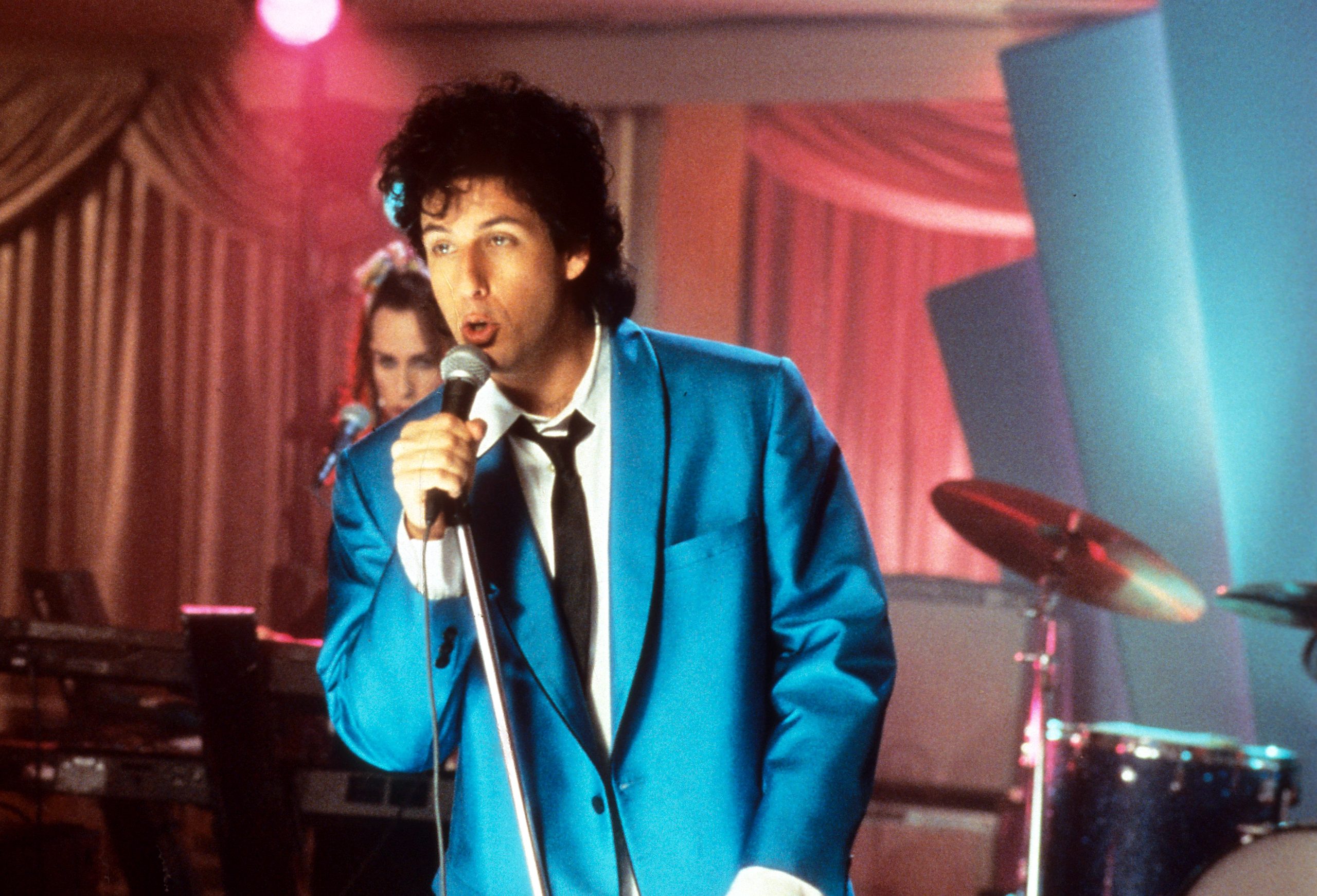 Adam Sandler in a bright blue suit jacket and black tie singing into a microphone on a stage.