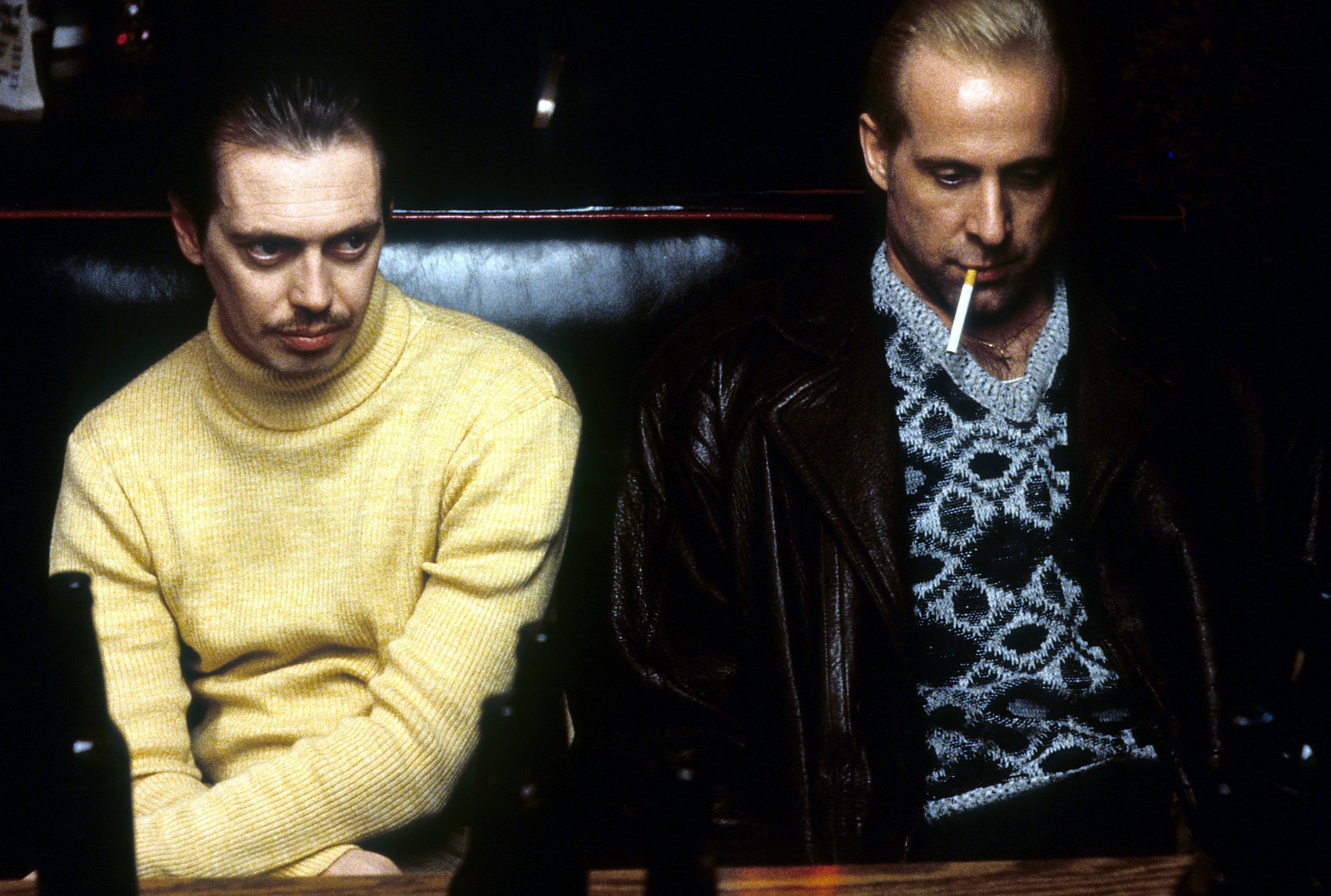Two men sit at a booth in a dimly lit bar, one in a yellow turtleneck and the other in a patterned sweater and leather jacket with a cigarette in his mouth.