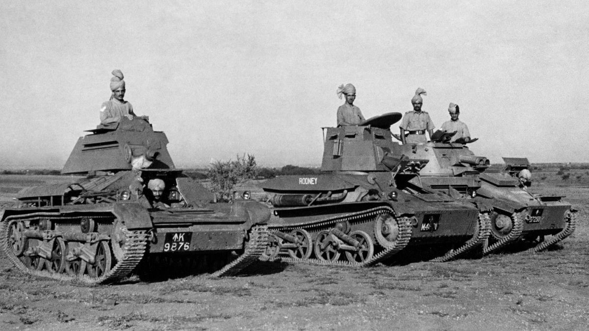 Four soldiers wearing turbans sit in small military tanks in a row on open ground. The tanks are lightly armored and have markings, with one labeled “Rodney.” The landscape is flat with sparse vegetation.