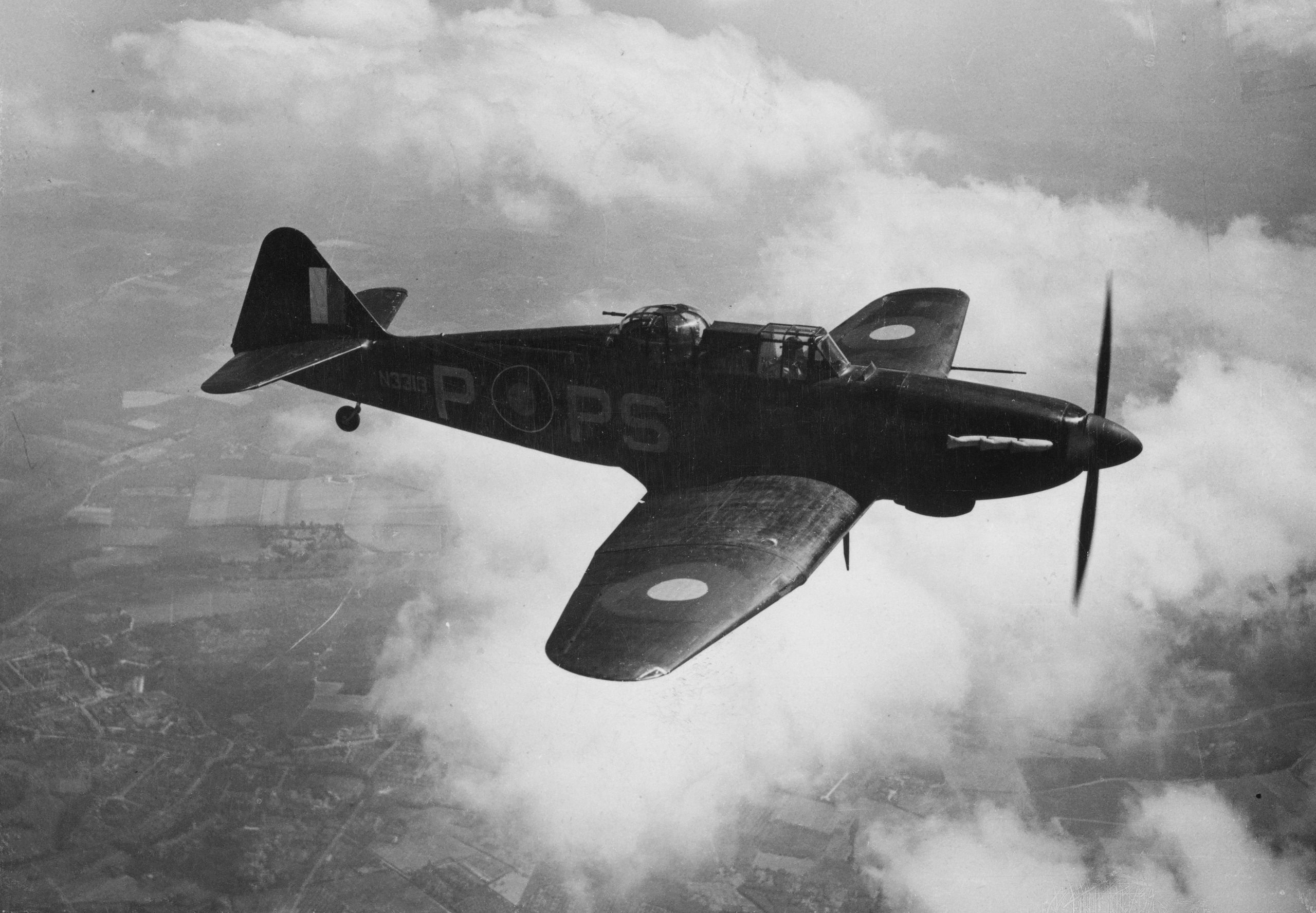 A World War II-era fighter aircraft with RAF markings flies above clouds over a landscape, seen from the side and slightly above.