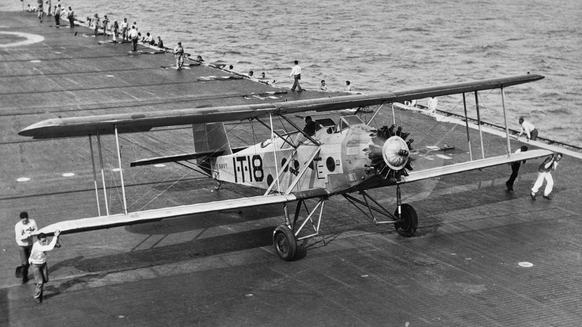 A vintage biplane sits on the deck of an aircraft carrier as crew members run alongside it; the ocean is visible in the background.