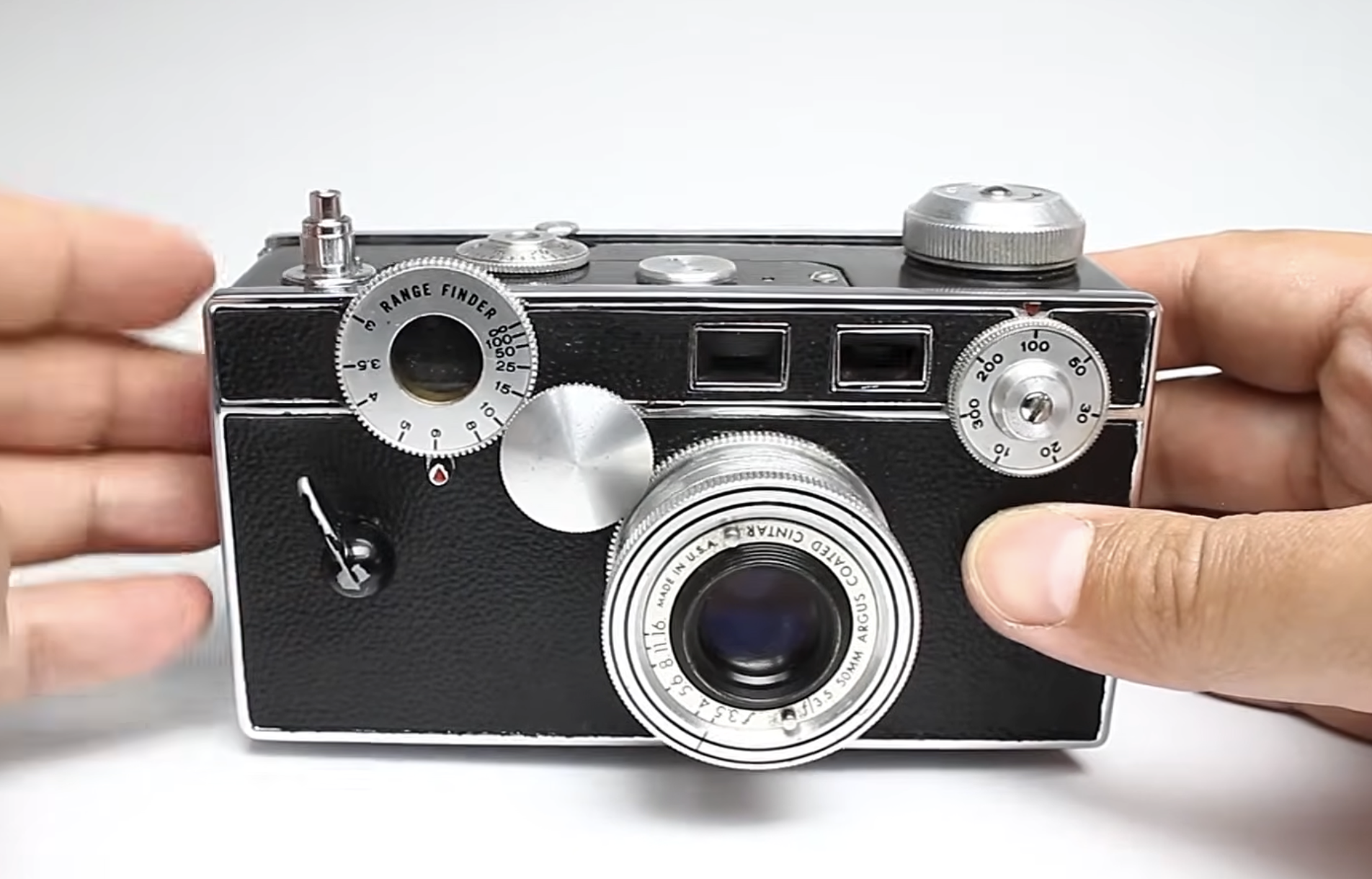 Hands hold a vintage Argus C3 rangefinder camera with a prominent lens and adjustment dials.
