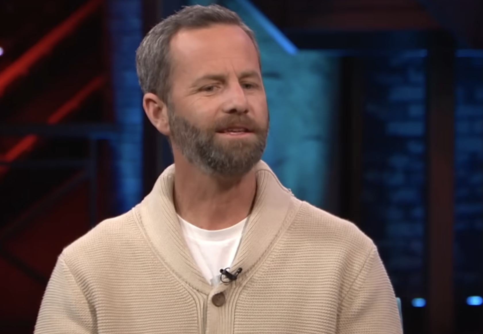 A middle-aged man with a short gray beard wearing a cream-colored cardigan over a white t-shirt.