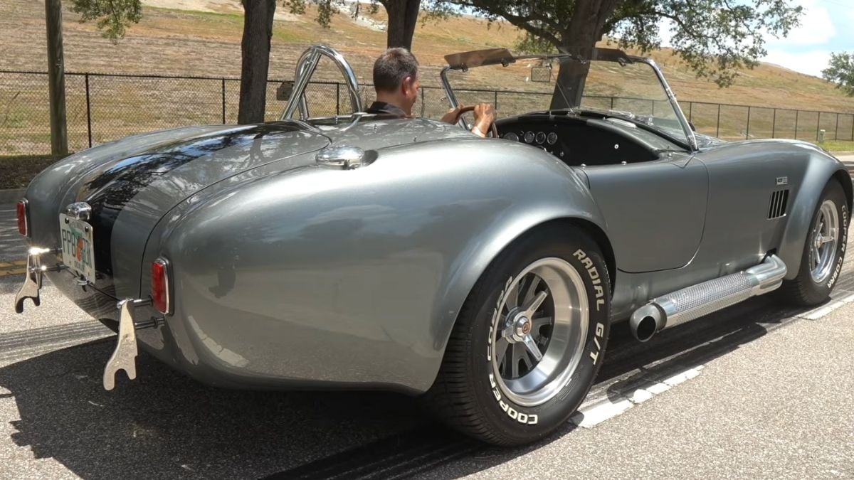 A classic silver convertible sports car with side exhaust pipes is parked on a road. A person is seated behind the wheel, and trees and a fence are visible in the background.