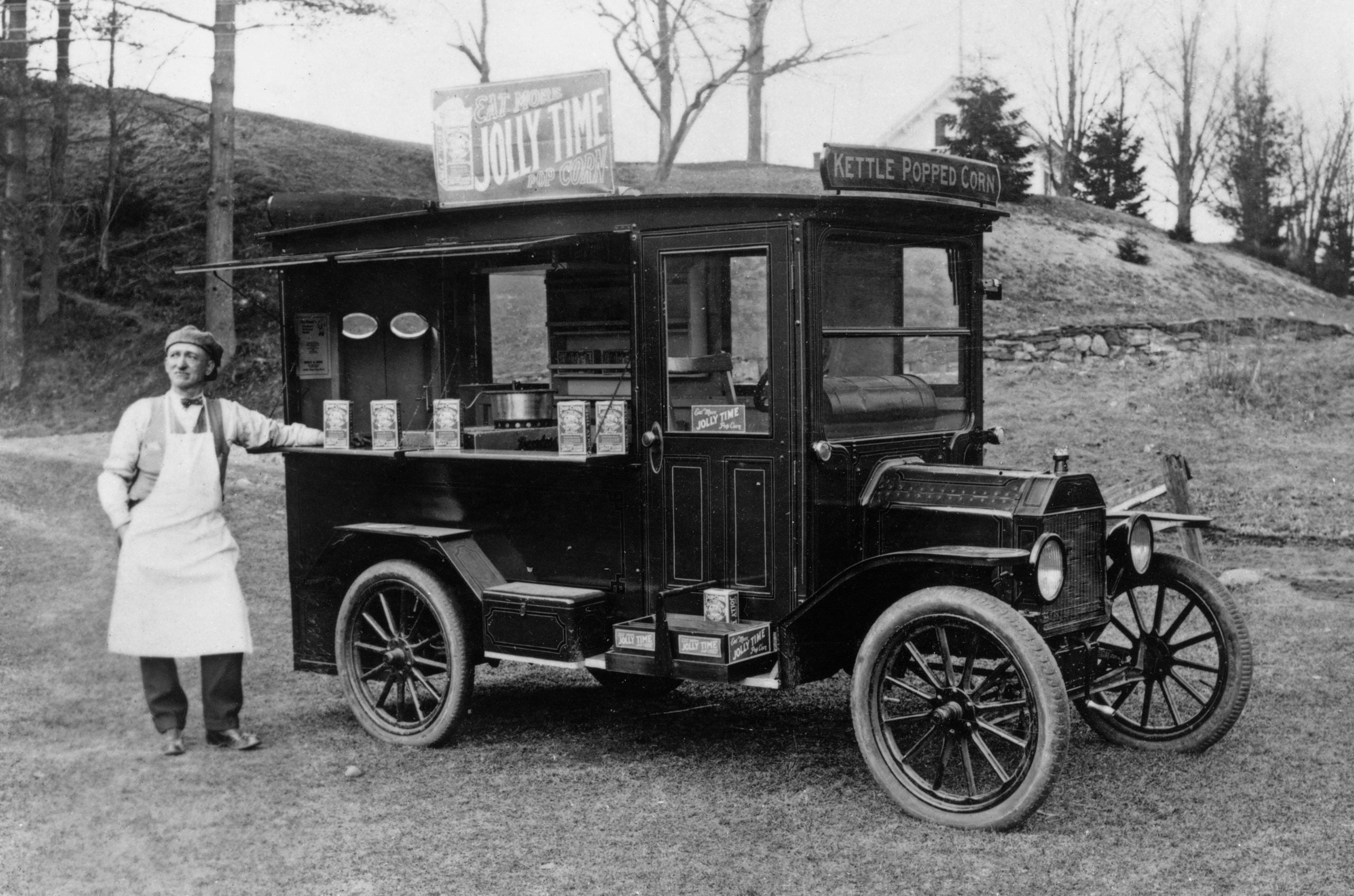 A man in an apron stands beside a vintage popcorn truck labeled “Jolly Time” and “Kettle Popped Corn” in a grassy outdoor area with trees and hills in the background.